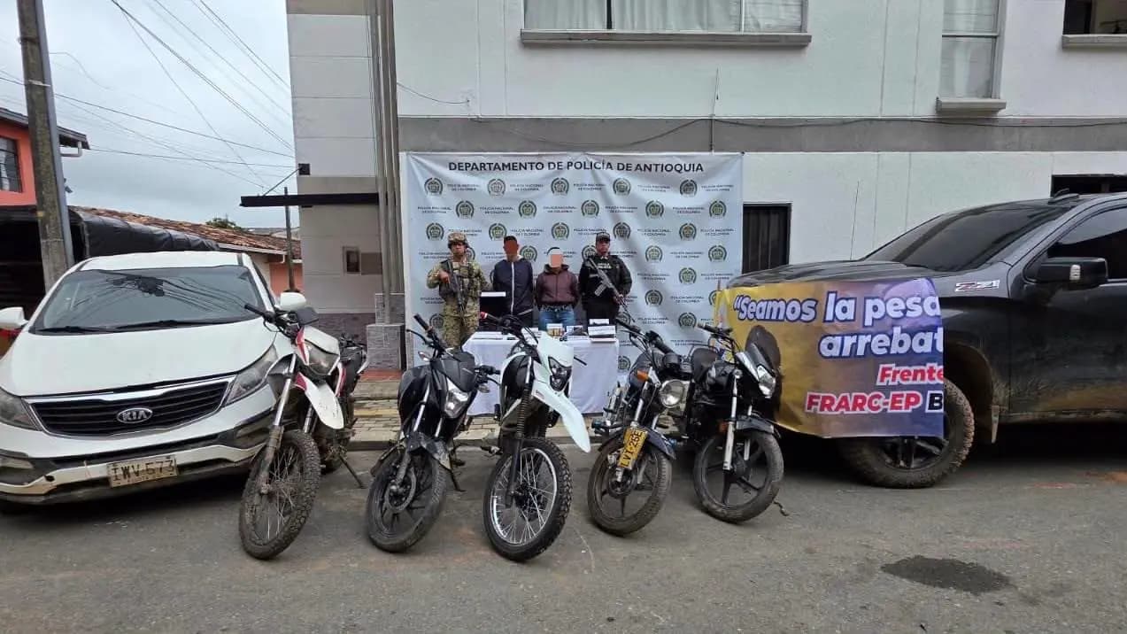 Los dos capturados en Gómez Plata, custodiados por un soldado y un policía, junto a las motos, las camionetas y una pancarta incautadas