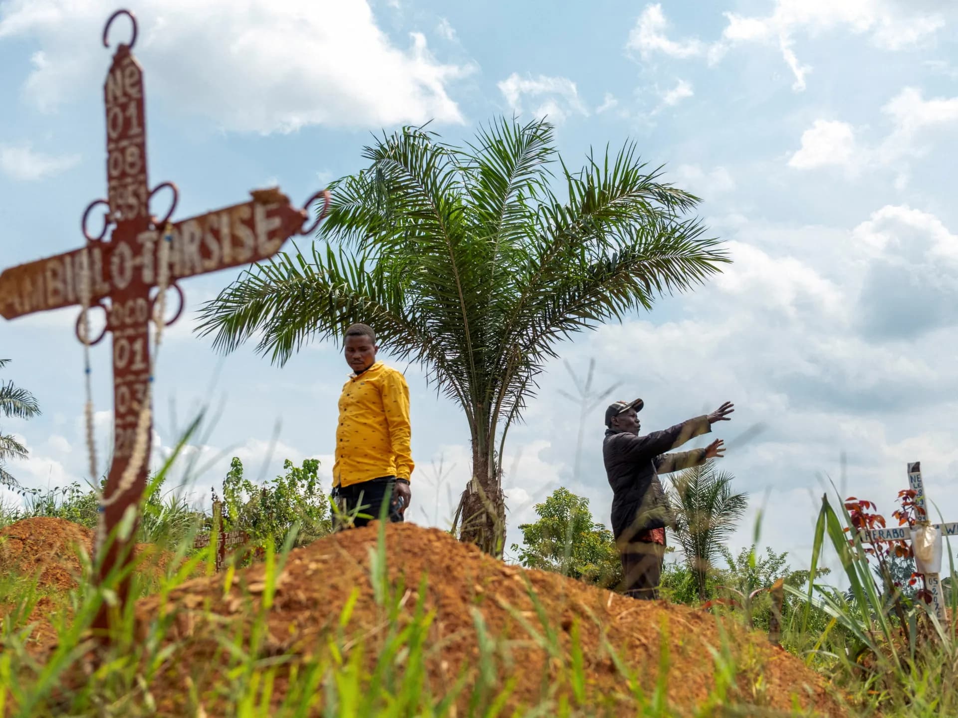 Dos hombres caminan entre cruces de madera pintadas en un cementerio de tierra roja en la provincia de Ituri, en el Congo
