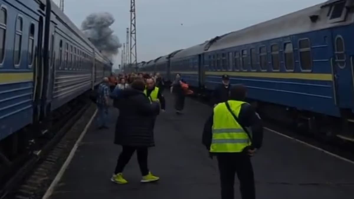 Pasajeros caminan entre dos trenes detenidos en una estación mientras una columna de humo gris se eleva al fondo