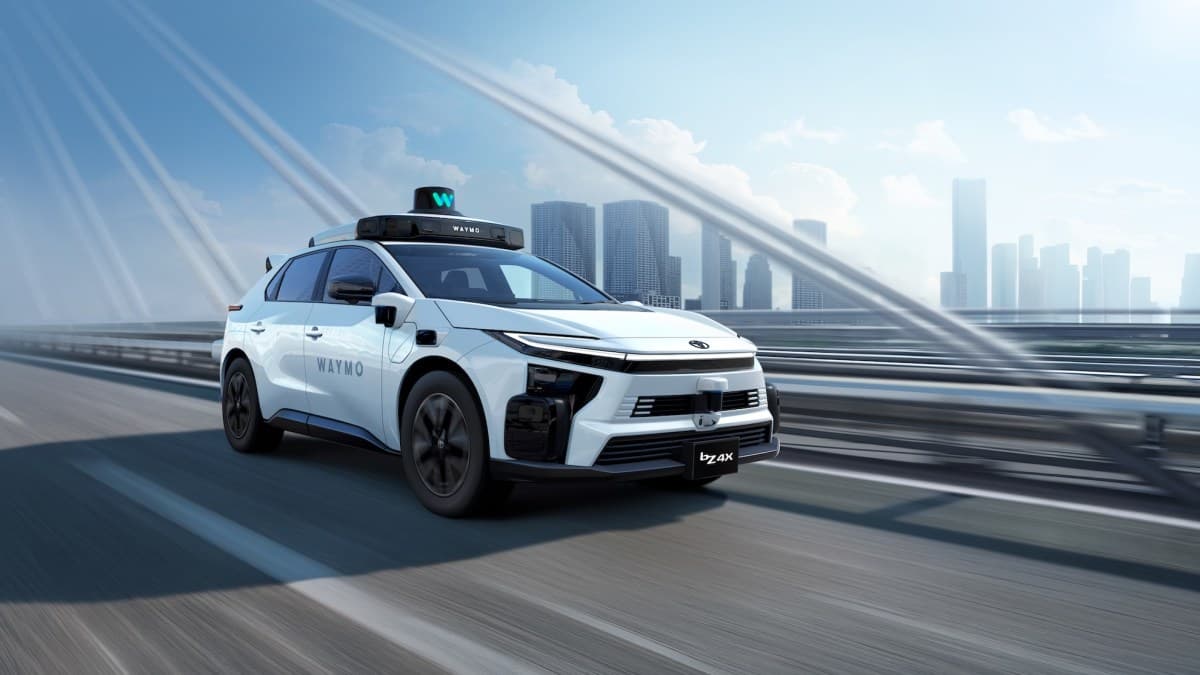 Toyota bZ4X blanco con los sensores de Waymo en el techo, en una autopista con una ciudad al fondo