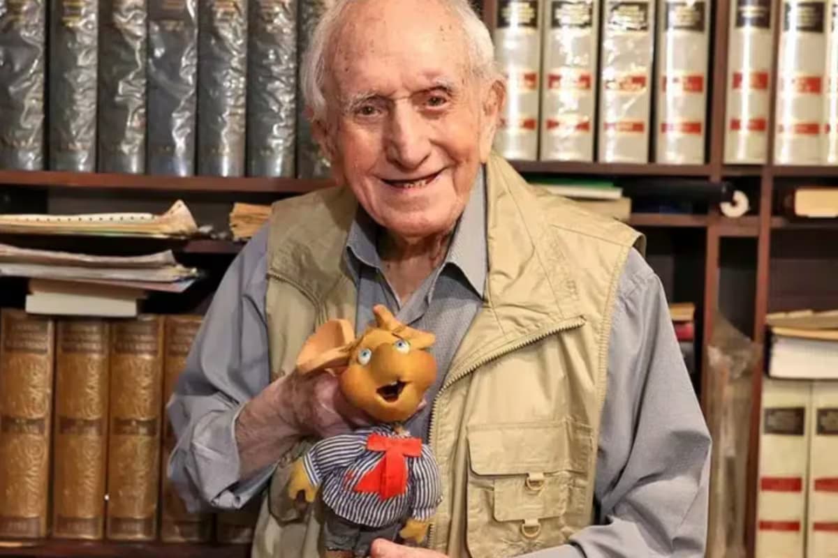 Peppino Mazzullo sonríe con un muñeco de Topo Gigio frente a una biblioteca