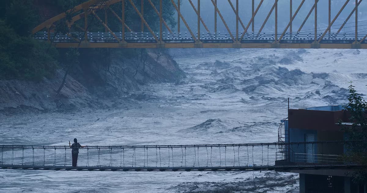 Un hombre cruza un puente colgante sobre un río crecido de aguas turbias que baja entre laderas de montaña, con un puente metálico amarillo al fondo