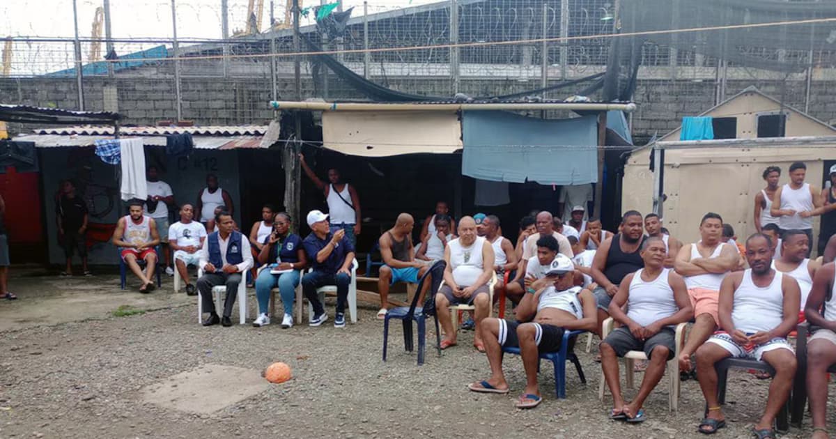 Decenas de detenidos sentados en sillas plásticas en un patio al aire libre bajo la vigilancia de funcionarios