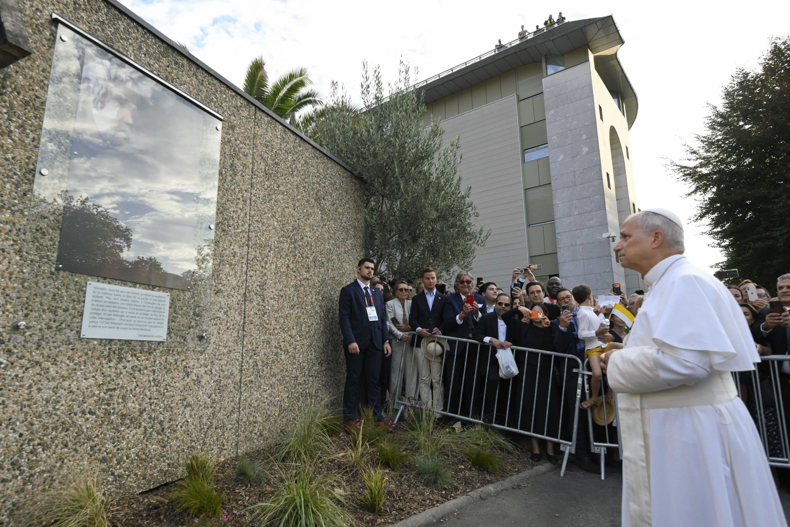 El papa León XIV, de blanco, ante un muro con una placa en Lourdes, mientras fieles lo observan tras una valla
