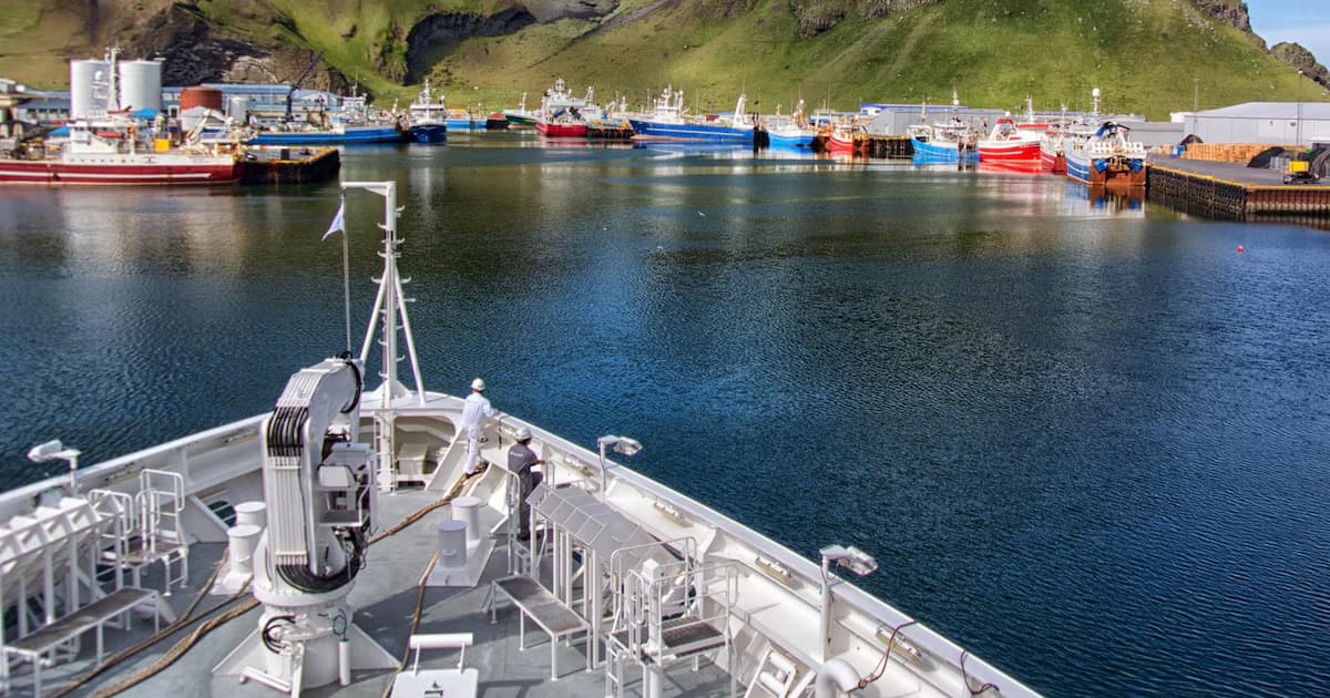 Barcos pesqueros de colores atracados en un puerto islandés al pie de una ladera verde, vistos desde la proa de un buque