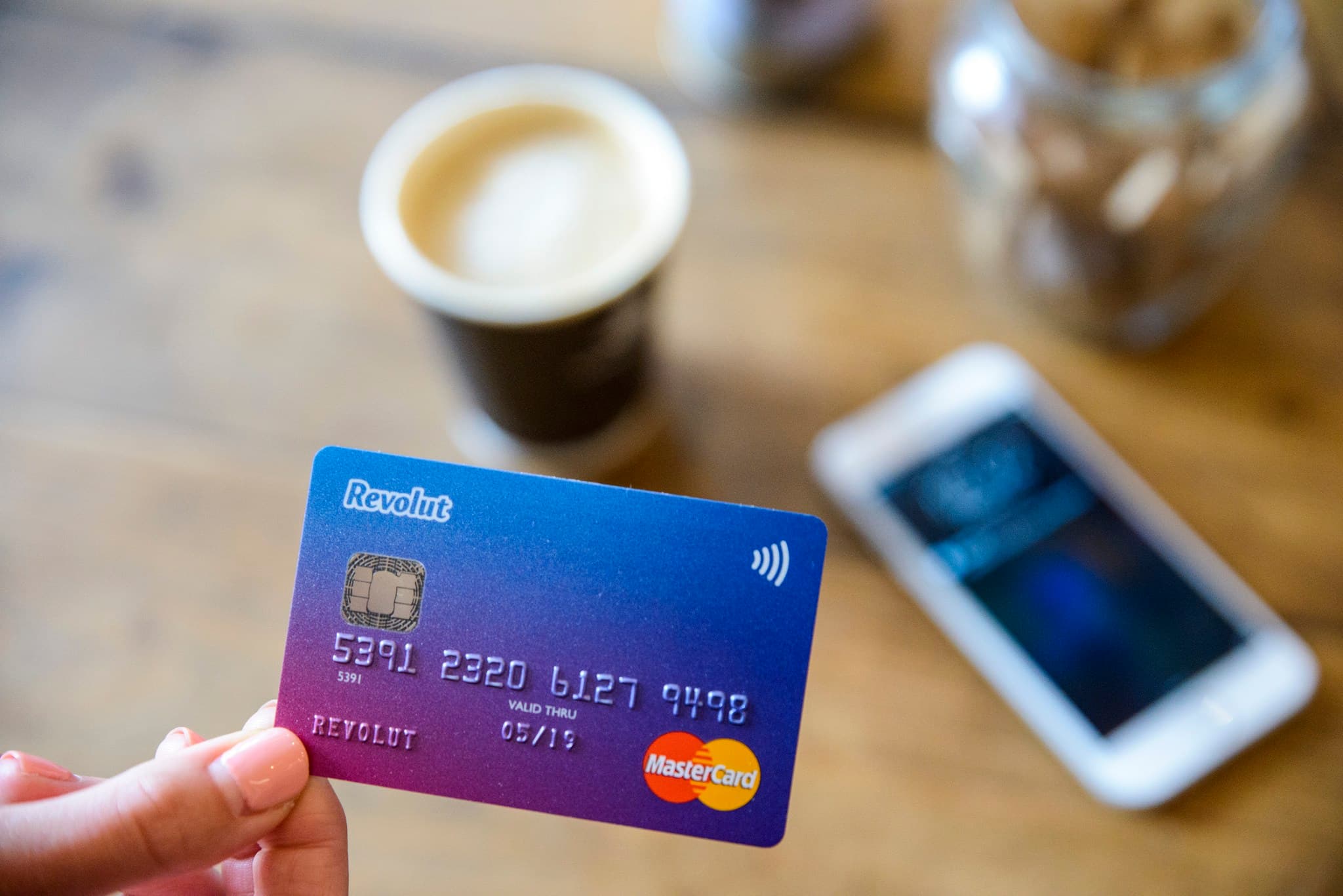 Una mano sostiene una tarjeta de Revolut frente a un café y un teléfono