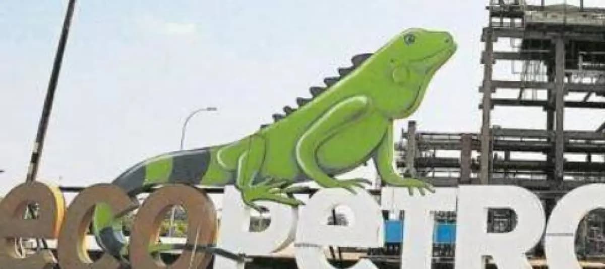 Letras corpóreas del nombre de Ecopetrol con una iguana verde encaramada, frente a las torres de una refinería