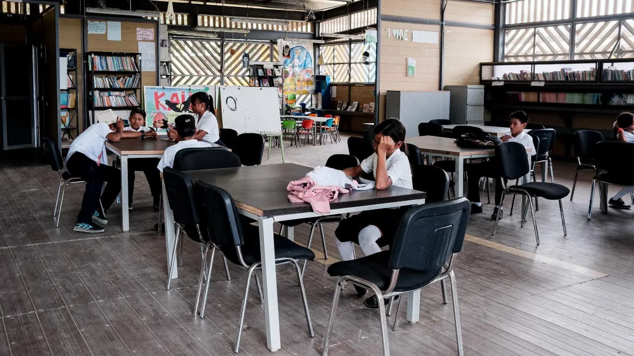 Estudiantes de uniforme blanco sentados en mesas de una biblioteca escolar con estanterías de libros al fondo