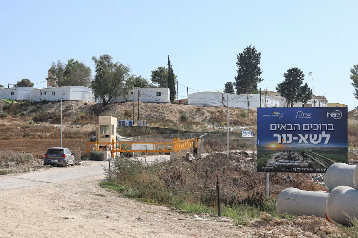 Entrada de un asentamiento israelí en Cisjordania, con una barrera amarilla, una garita y viviendas prefabricadas blancas en la loma