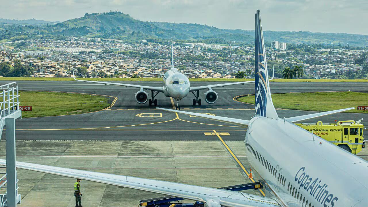 Un avión rueda por la pista del aeropuerto Matecaña con Pereira y las montañas al fondo, junto a otra aeronave estacionada en plataforma