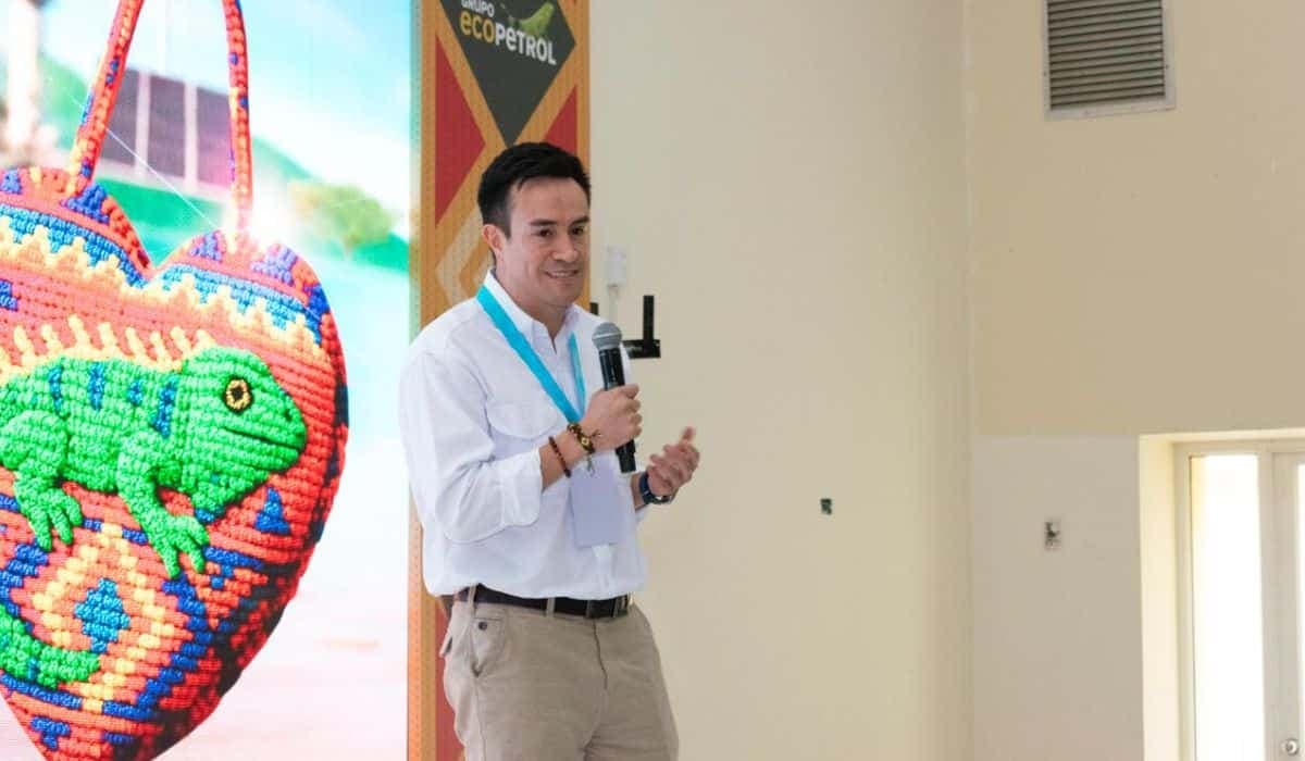 Bayron Triana habla con un micrófono en la mano durante un evento empresarial, junto a una pantalla con una mochila artesanal