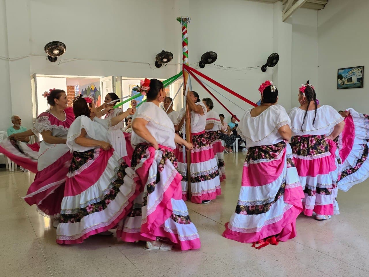 Un grupo de mujeres con falda larga de flores y blusa blanca baila alrededor de un palo de cintas en un salón comunitario