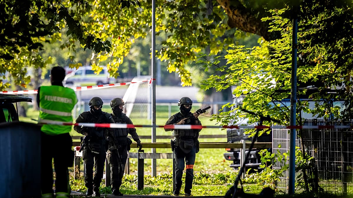 Tres agentes con equipo táctico y armas largas caminan por un prado acordonado con cinta roja y blanca, junto a un bombero de chaleco reflectivo