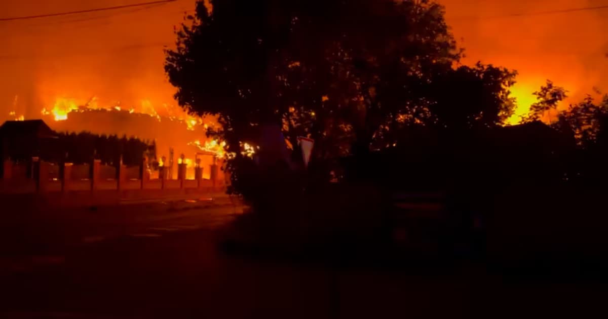 Un incendio de grandes proporciones tiñe de naranja el cielo nocturno detrás de las siluetas de árboles y de una construcción baja