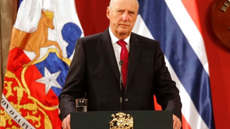 El rey Harald V de Noruega, de traje oscuro y corbata roja, de pie ante un atril con la bandera noruega detrás