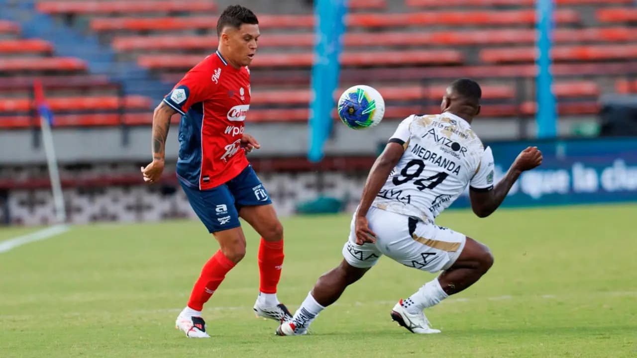 Un jugador del Medellín con camiseta roja y azul controla el balón ante la marca de un rival de blanco en el césped del Atanasio Girardot