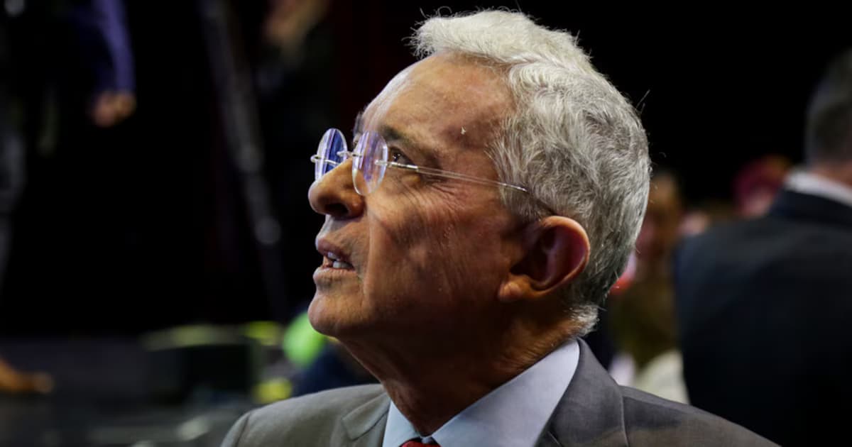 Álvaro Uribe Vélez, de perfil, con gafas de montura fina y traje gris, en un recinto de techo oscuro