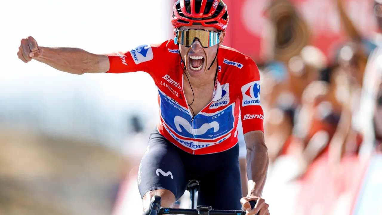 Un ciclista con maillot rojo de líder y gafas doradas cruza la meta con el brazo extendido y la boca abierta en un grito de celebración