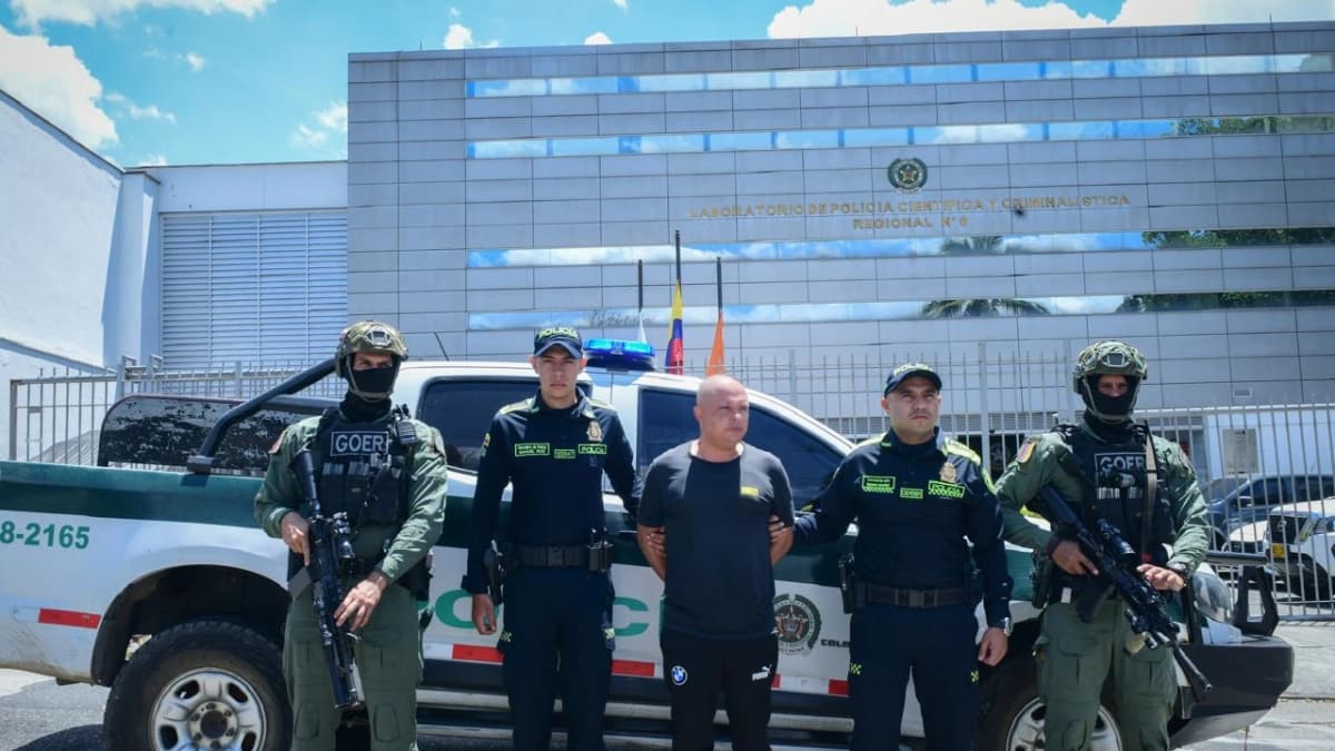 Un hombre custodiado por cuatro policías y miembros de un grupo de operaciones especiales frente a una camioneta de la Policía y al laboratorio de criminalística