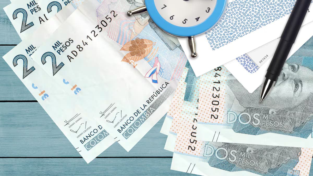 Billetes de dos mil pesos colombianos extendidos sobre una mesa de madera junto a un reloj y un bolígrafo