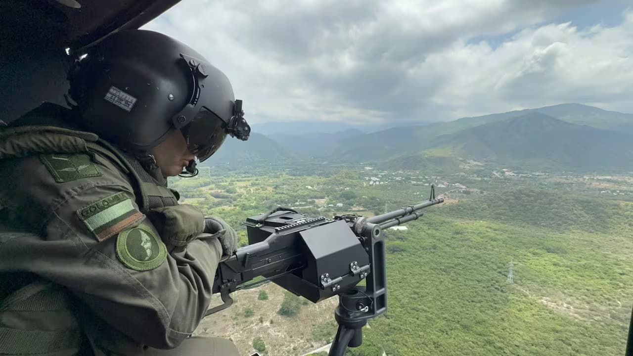 Un militar con casco de vuelo opera una ametralladora desde la puerta de un helicóptero sobre una zona montañosa
