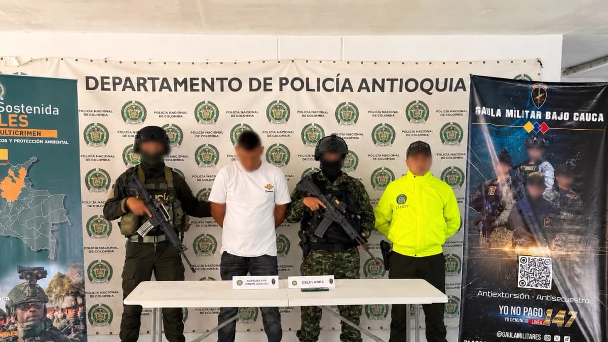 Un hombre detenido, con el rostro cubierto, custodiado por uniformados frente a un pendón del Departamento de Policía Antioquia