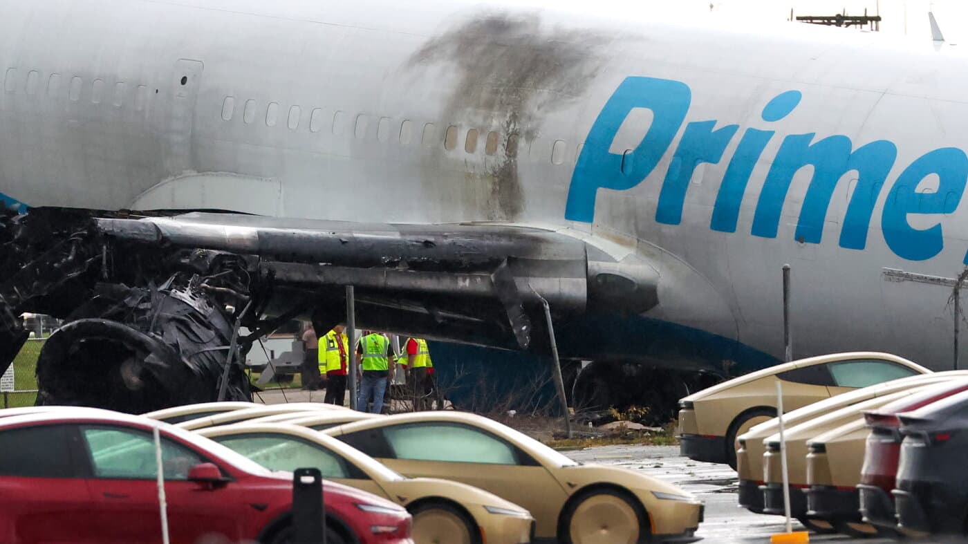 Operarios junto al fuselaje quemado de un avión de carga de Amazon tras salirse de la pista