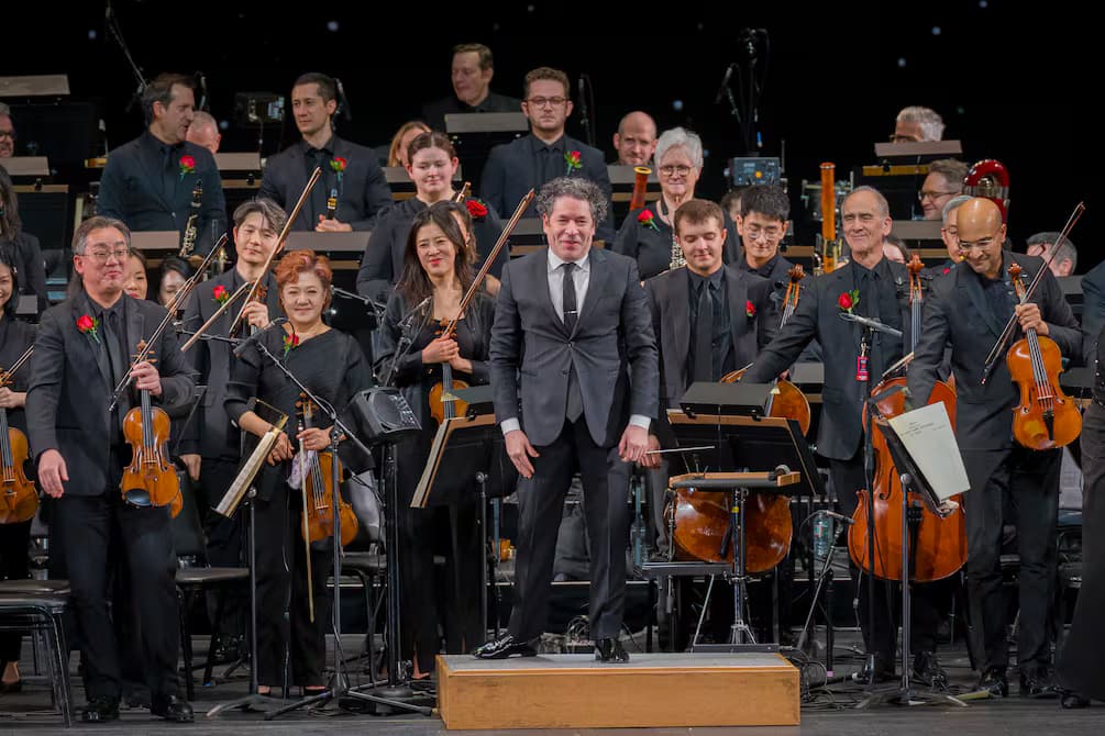 Gustavo Dudamel de pie en el podio saluda al público junto a los músicos de la orquesta, que aplauden con sus instrumentos en la mano
