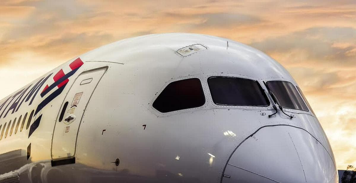 Morro de un avión de LATAM estacionado en la plataforma de un aeropuerto con el cielo anaranjado del atardecer detrás