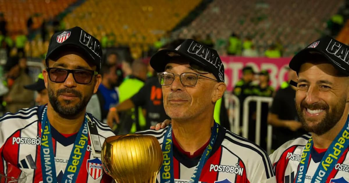 Alfredo Arias, con camiseta de Junior y gorra de campeón, posa junto al trofeo de la Liga BetPlay acompañado por dos miembros de su cuerpo técnico