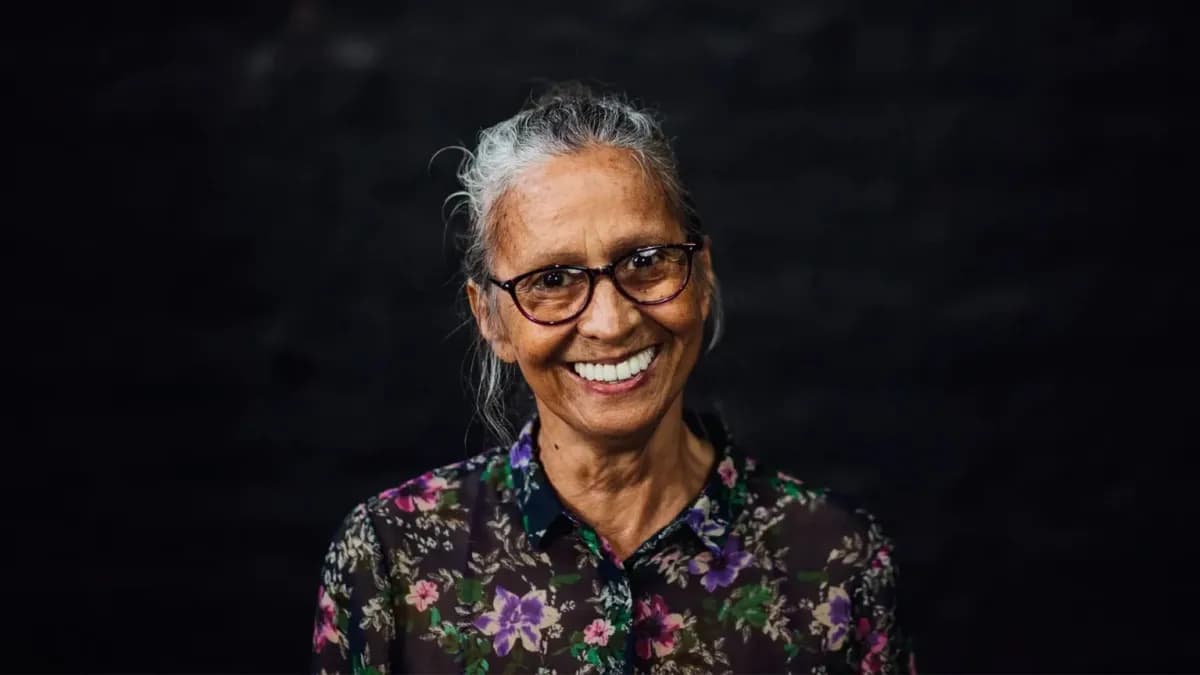 Retrato de una mujer mayor de pelo cano recogido, gafas de pasta y camisa estampada de flores, sonriendo sobre un fondo oscuro