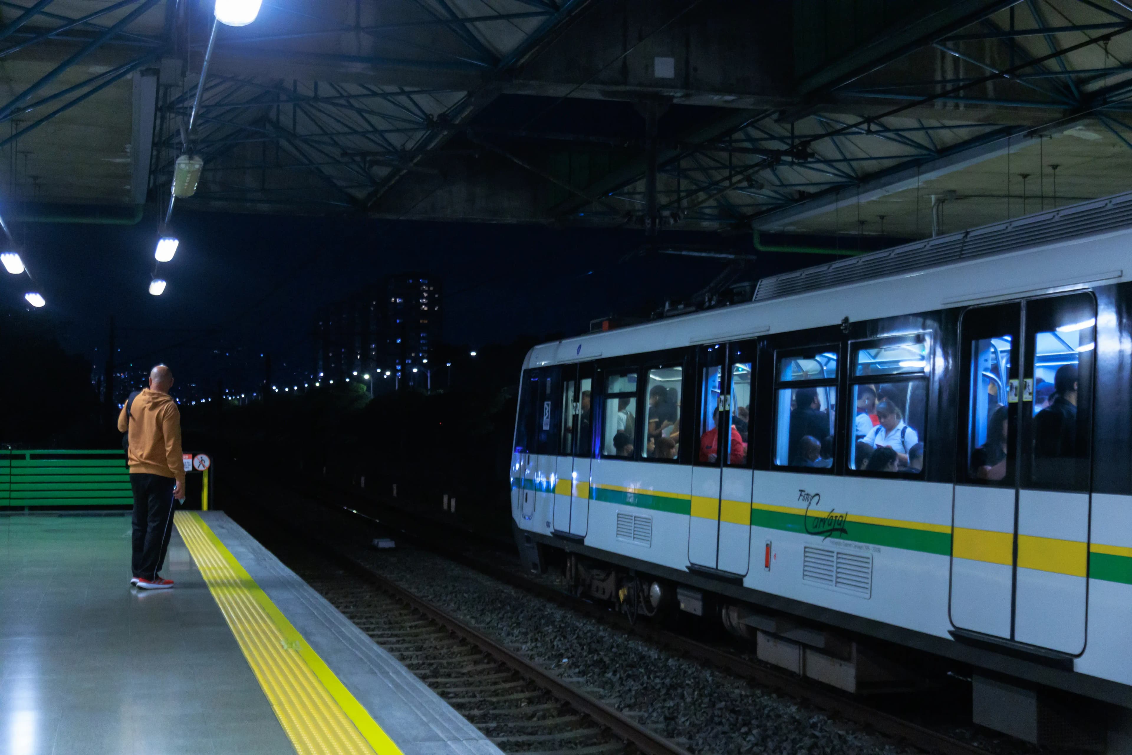 Un tren del metro de Medellín llega de noche a una estación mientras un pasajero espera en la plataforma