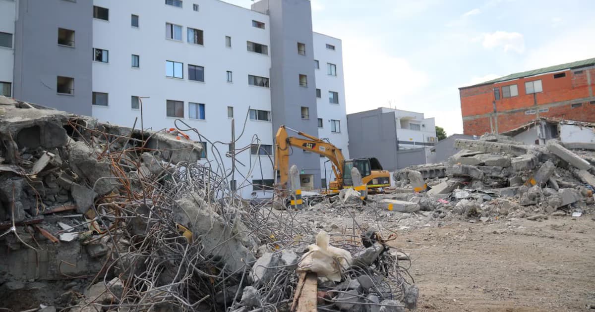 Una retroexcavadora trabaja entre los escombros de un edificio derrumbado, junto a un bloque de apartamentos en pie