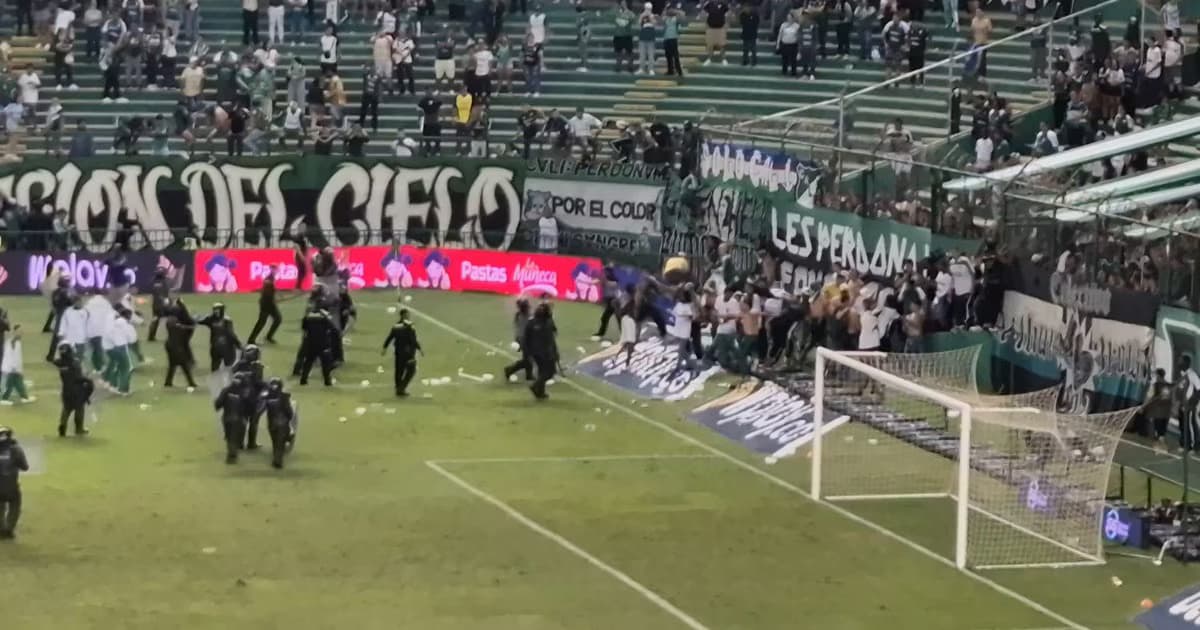 Policías antidisturbios corren por el césped de un estadio junto a la tribuna mientras un grupo de aficionados se agolpa sobre una valla derribada