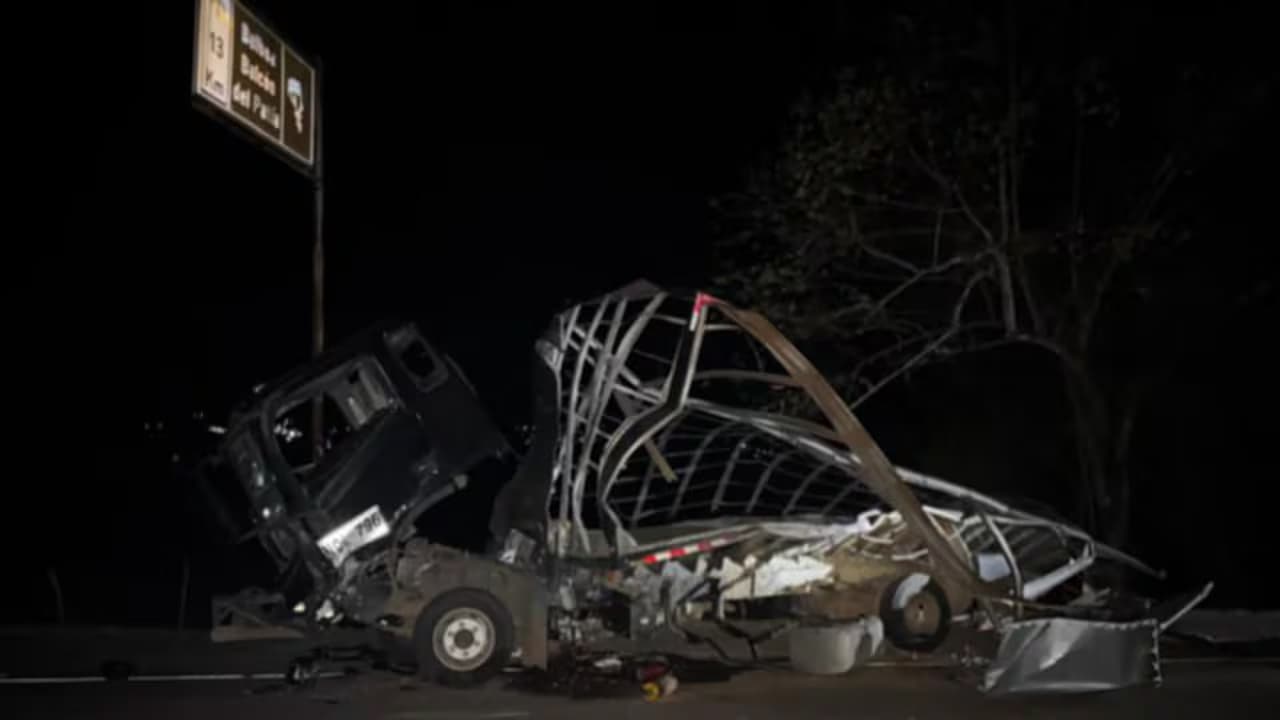 Restos calcinados y retorcidos de un camión sobre una carretera de noche, junto a una valla de señalización vial