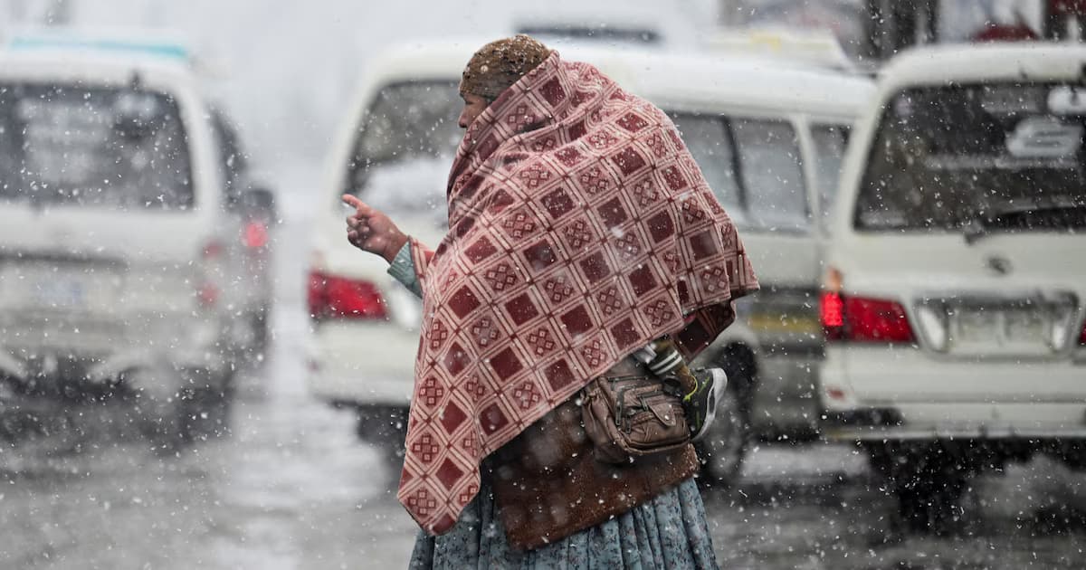 Una mujer cubierta con una manta cruza una calle bajo la nieve, entre vehículos detenidos