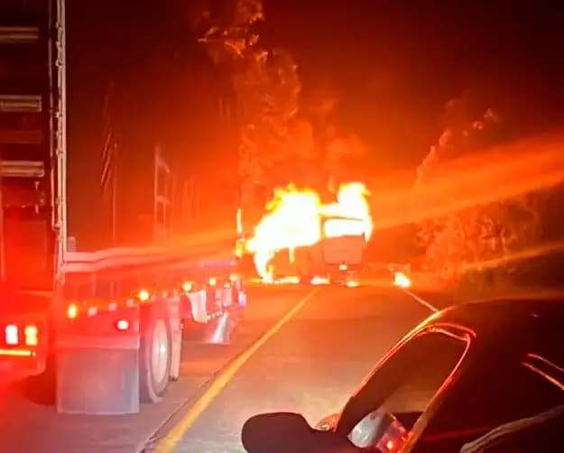 Un bus intermunicipal envuelto en llamas en plena carretera de noche, visto desde un vehículo detenido detrás de un camión