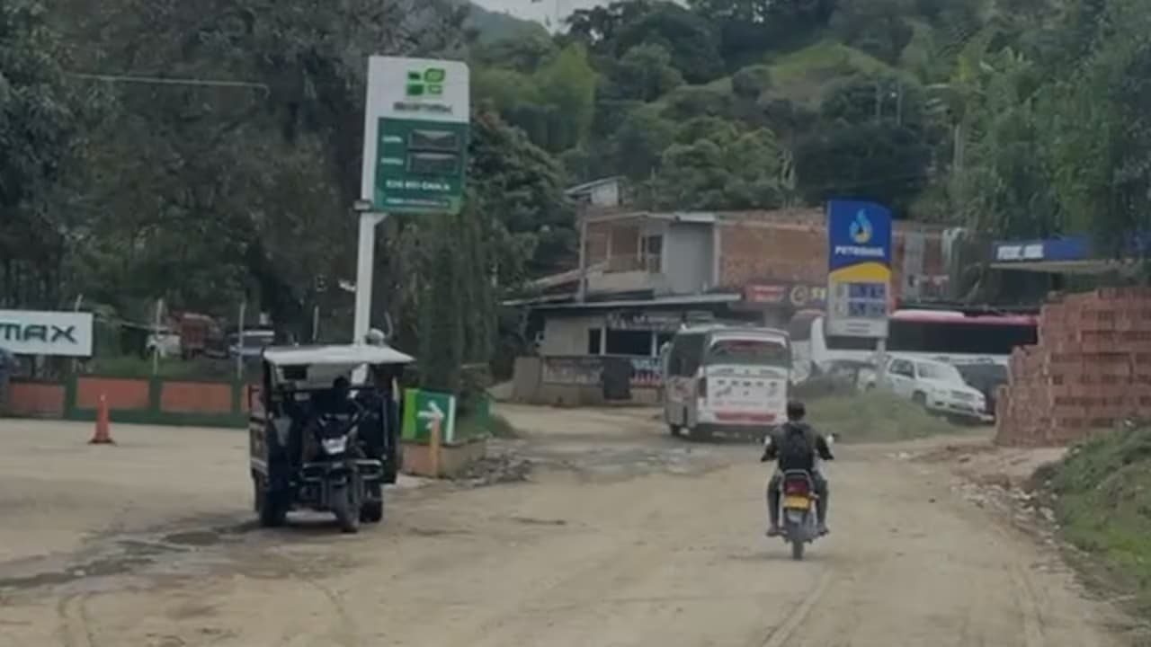 Una calle destapada de un casco urbano del norte del Cauca, con un motocarro, una moto y un bus de escalera junto a una estación de gasolina y casas de ladrillo en la ladera