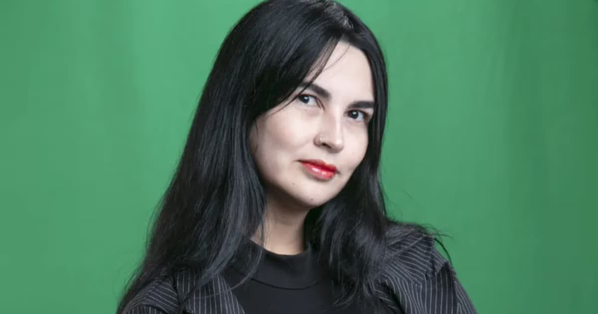 Retrato de Hannah Escobar, de cabello largo y oscuro y saco de rayas, mirando a la cámara sobre un fondo verde