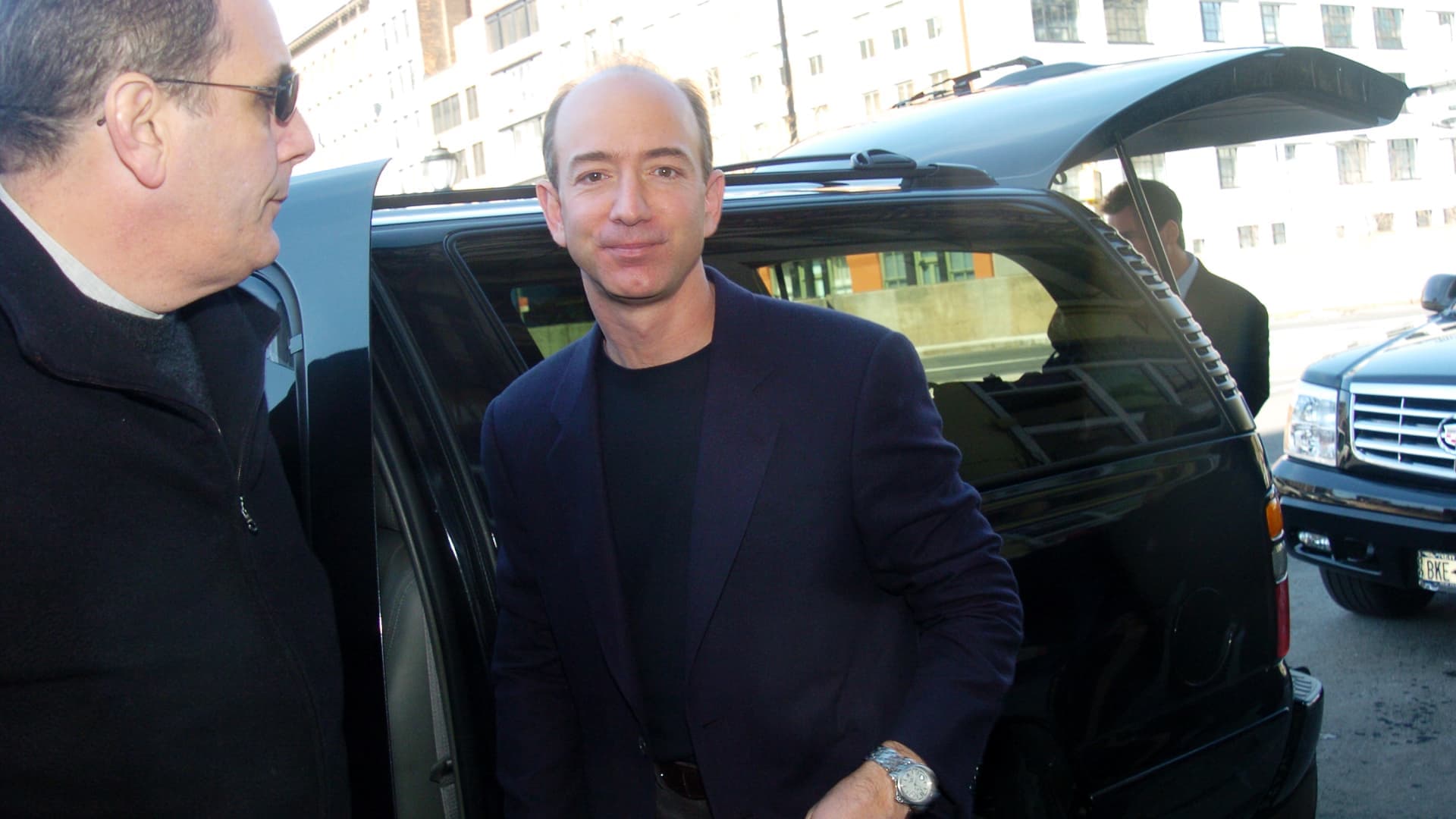 Jeff Bezos, de saco oscuro, sonríe al bajar de una camioneta negra en una calle de la ciudad