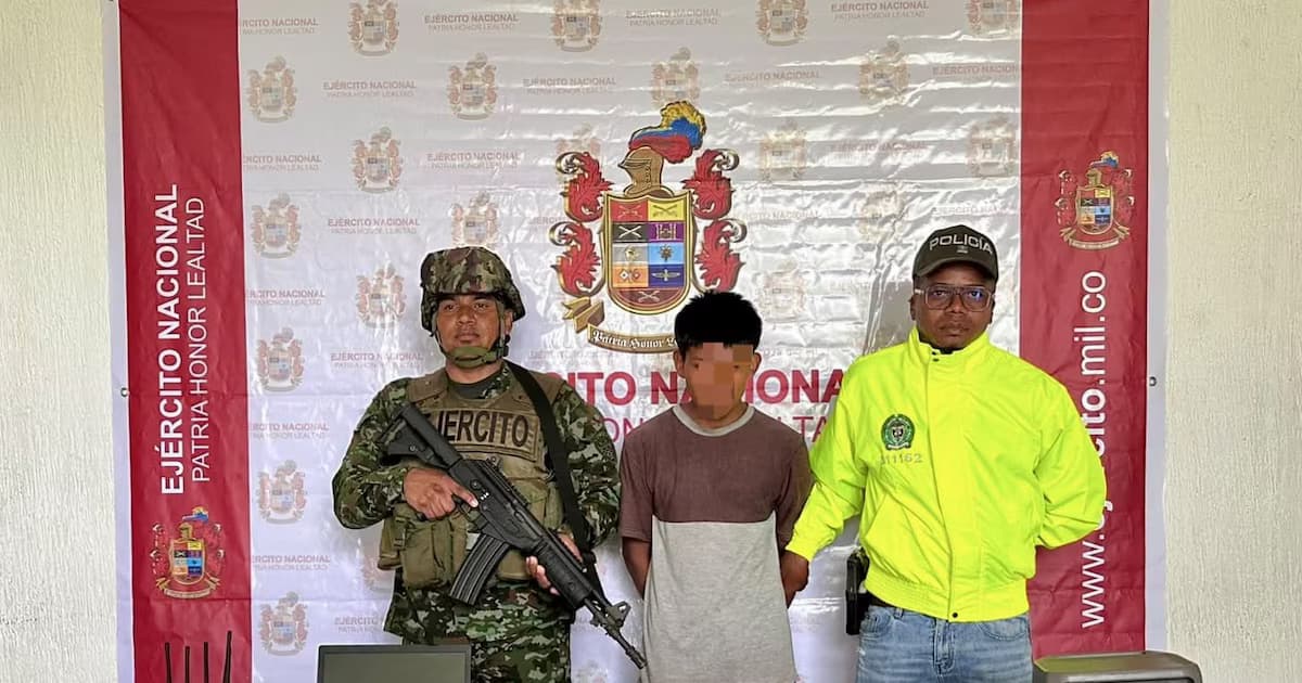 Un soldado y un policía custodian a alias el Tuerto, con el rostro difuminado, frente a un pendón del Ejército