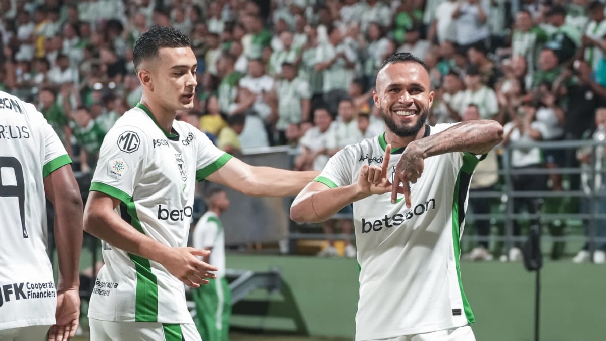 Dos futbolistas de Atlético Nacional con la camiseta blanca y verde celebran un gol haciendo un gesto con las manos frente a la tribuna llena