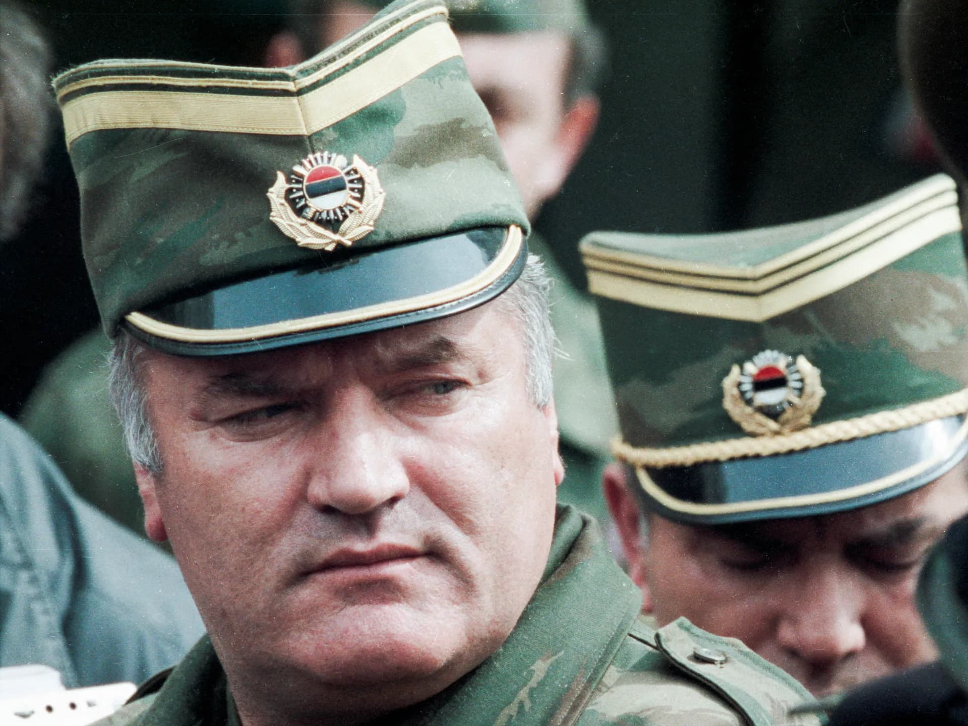 Ratko Mladic, con uniforme de campaña y gorra militar con la escarapela serbia, mira hacia abajo rodeado de oficiales durante la guerra de Bosnia