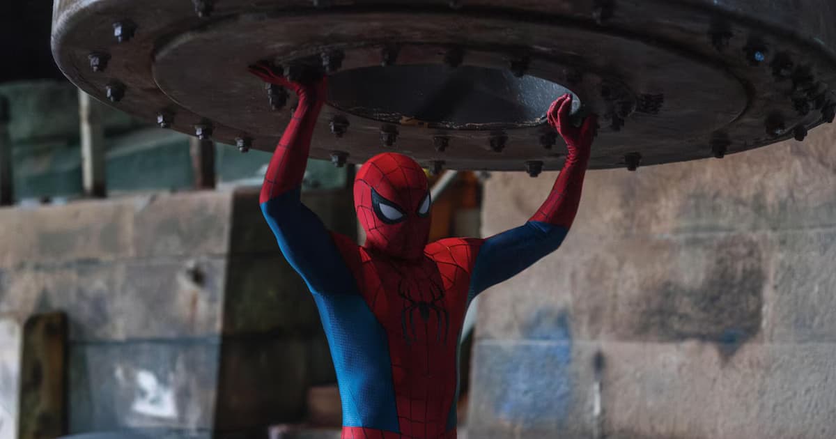 Spider-Man, con el traje rojo y azul, levanta con ambos brazos una gran tapa metálica circular dentro de una nave industrial