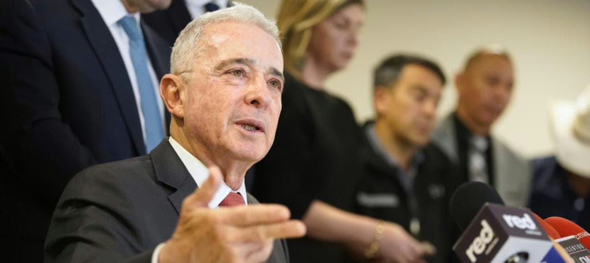 El expresidente Álvaro Uribe habla ante un grupo de micrófonos con la mano extendida, rodeado de acompañantes