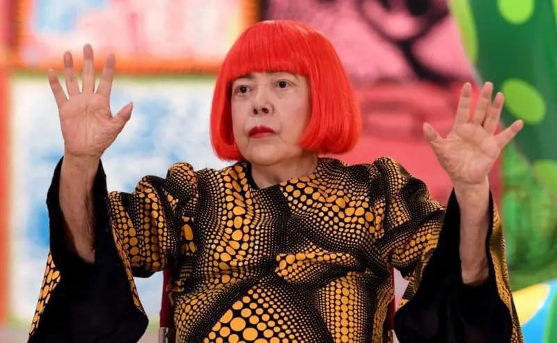 Yayoi Kusama, con peluca roja y un vestido negro cubierto de lunares amarillos, levanta las dos manos abiertas frente a sus cuadros de colores