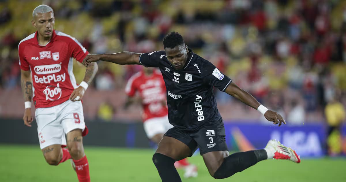 Un jugador del América de Cali con camiseta negra remata ante la marca de un rival de camiseta roja