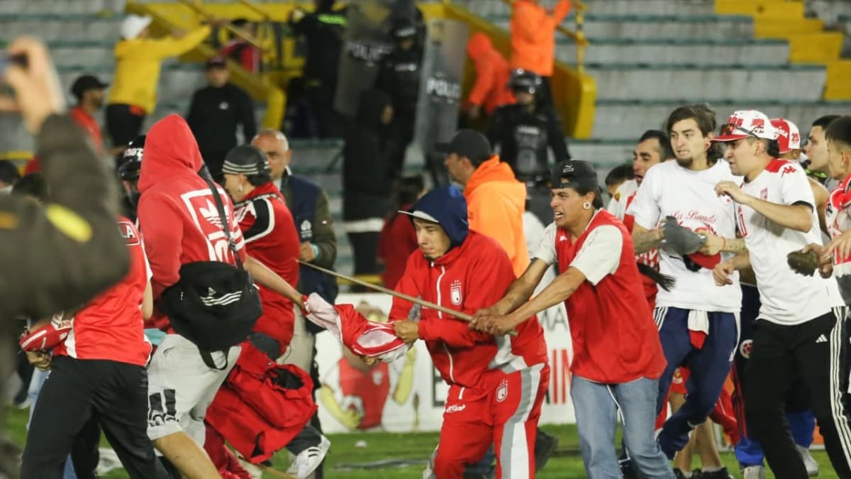 Hinchas de Santa Fe y América forcejean por una bandera sobre el terreno de juego de El Campín, con policías antimotines al fondo