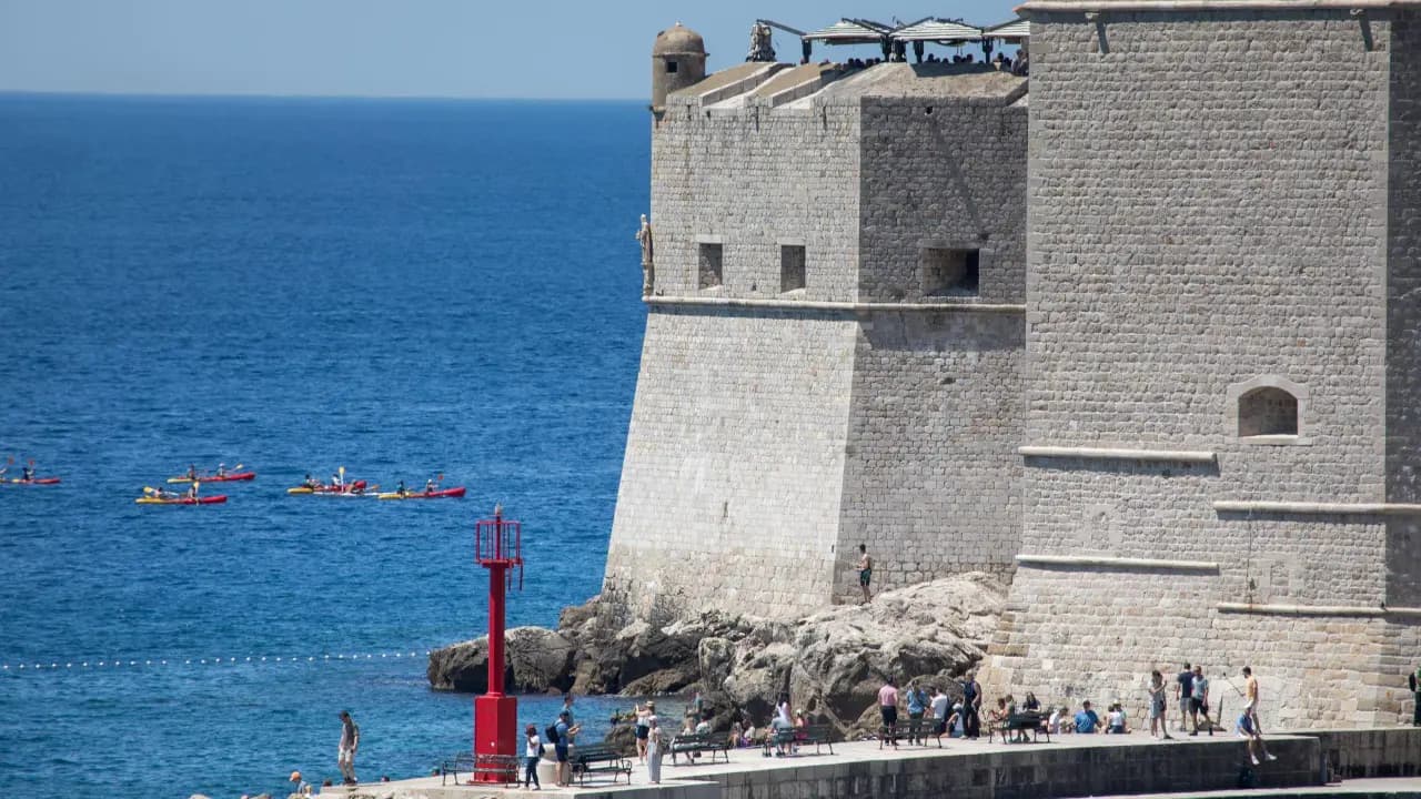 Bañistas y kayaks en un mar de aguas azules junto a una muralla de piedra en un día despejado de verano