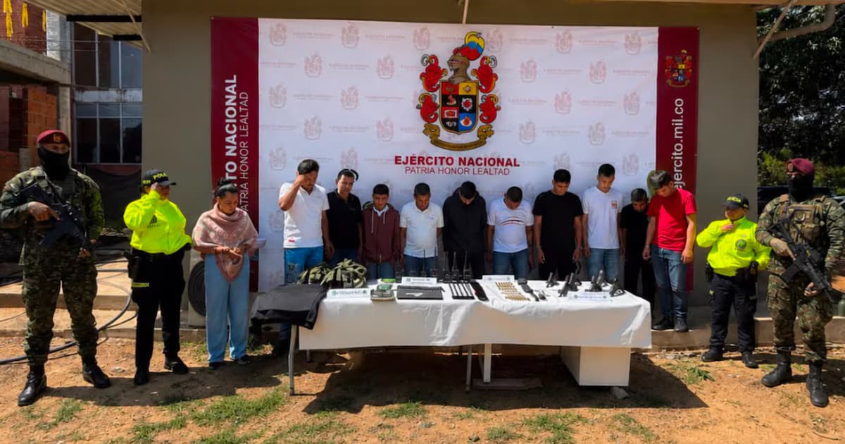 Once personas capturadas, custodiadas por militares y policías, frente a una mesa con armas incautadas y un pendón del Ejército Nacional